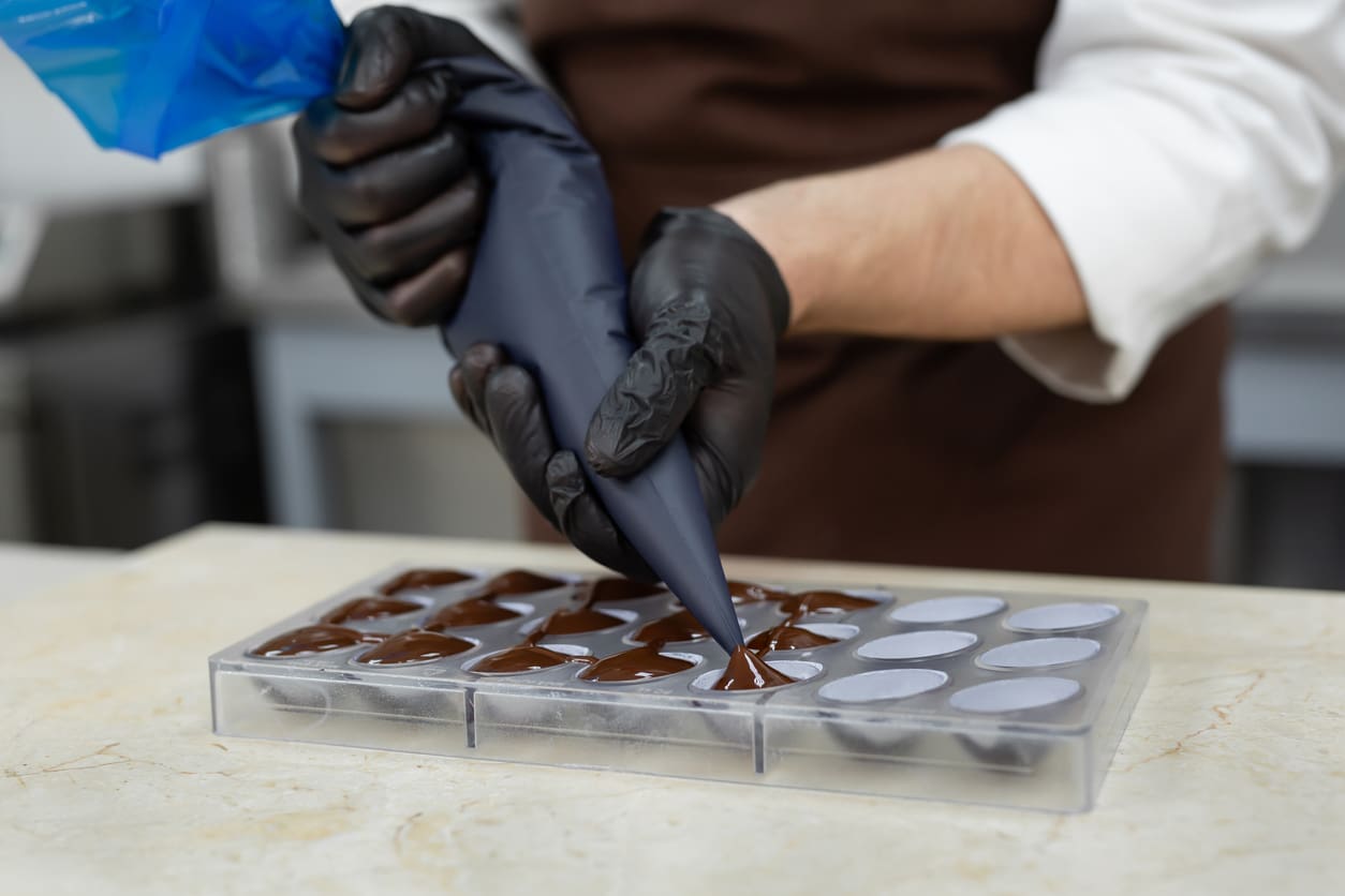 Close-up of the chocolatier's hands pouring chocolate into the molds. The chef's pastry bag pours hot melted chocolate into a silicone mold.